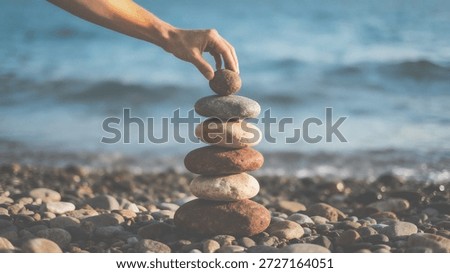 A person's hand carefully balances a stack of smooth stones on a rocky beach with blurred ocean waves in the background, conveying a sense of serenity and calmness in a natural setting with various sh
