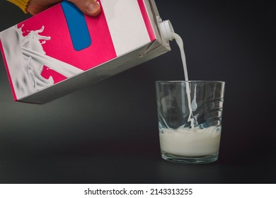 A person's hand holding a carton of delicious fresh milk and pouring it into a glass. Image of a stream of milk filling a small glass on a black background. - Shutterstock ID 2143313255