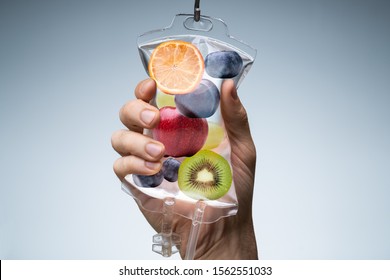 Person's Hand Holding Saline Bag Filled With Various Fruit Slices Against Grey Backdrop