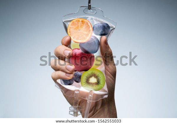 Person\'s Hand Holding Saline Bag Filled\
With Various Fruit Slices Against Grey\
Backdrop