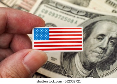 Persons Hand Holds American Flag Badge (USA Flag Pin) Against Heap Of Dollar Bills. Close Up.