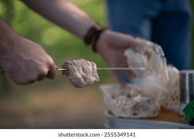 persons hand holds a skewer with raw meat over a barbecue. The background is blurred - Powered by Shutterstock