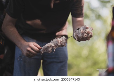 persons hand holds a skewer with raw meat over a barbecue. The background is blurred - Powered by Shutterstock