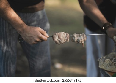 persons hand holds a skewer with raw meat over a barbecue. The background is blurred - Powered by Shutterstock