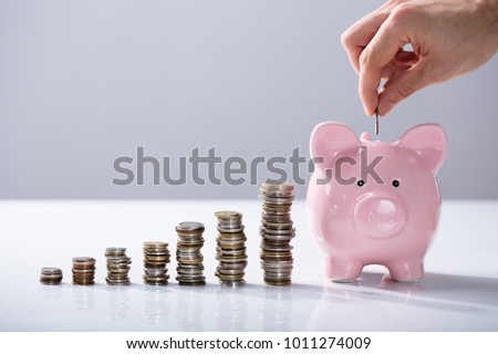 Person's Hand Inserting Coin In The Piggybank With Increasing Coins Stack On Desk