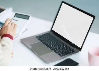 Person's hand interacting with a calculator, analyzing financial data and reviewing accounting figures while working remotely at a desk with a laptop displaying a blank screen