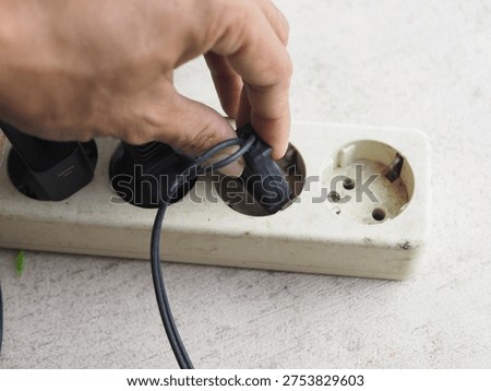Person's hand plugging a black electrical cable into a white power strip, connecting to a power outlet