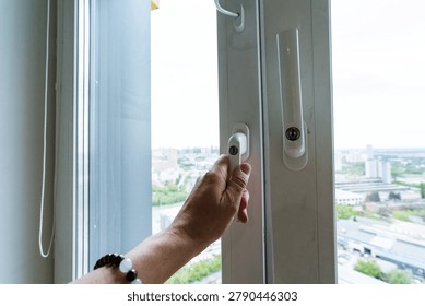 A persons hand pulls a window latch from indoors, standing near a glass pane with sheer curtains. Bright daylight streams in, revealing an airy office-like setting and distant buildings outside, suggesting ventilation, fresh air, and a calm city view