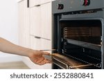 Persons hand reaches into an open oven, pulling out a metal grill grid. Woman cleaning kitchen appliances and household equipment. Concepts of chores and housework