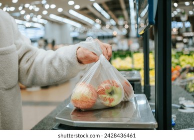 A person's hand is shown placing a plastic bag of fresh apples on a digital scale in the produce section of a grocery store, ensuring the weight is correct for checkout. - Powered by Shutterstock