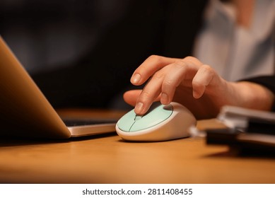 Persons hand using a mint green computer mouse on a wooden desk next to a laptop, close-up view