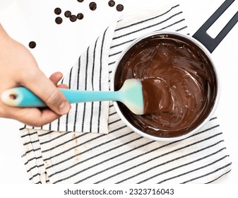 A person's hand using spatula stir to melt chocolate in saucepan before bakes cake and cookies. Pasty and bakery chef mixing ingredients in pot, prepare for homemade dessert. Cooking object on napkin.