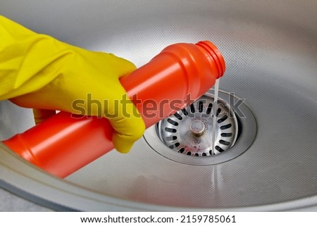 Person's hand in a yellow rubber glove pours pipe cleaner down the drain of a metal kitchen sink