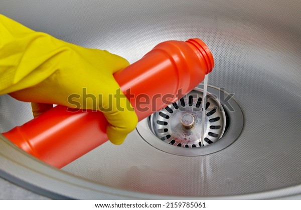 Person\'s hand in a yellow rubber\
glove pours pipe cleaner down the drain of a metal kitchen\
sink