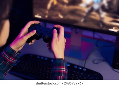 A persons hands hold a black video game controller, with their fingers resting on the buttons, playing a video game on a computer monitor, which is lit up with a colorful screen displaying the action. - Powered by Shutterstock