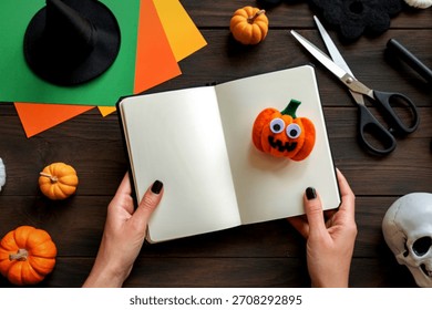 A person's hands hold an open notebook with a handmade felt pumpkin. Craft supplies are scattered around, ready for Halloween projects.