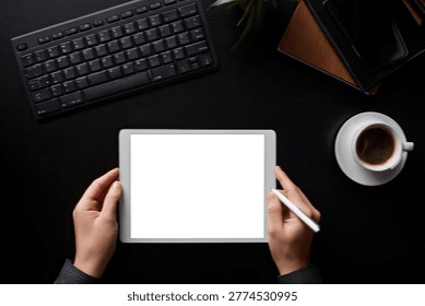 A person's hands hold a white tablet with a blank screen, a stylus in the right hand, on a black desk with a keyboard and coffee cup.