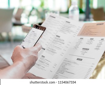 Person's Hands Holding Menu And Mobile Phone In Restaurant. A Foreign Man In Cafe Ordering Food Using Word Translation Program While Sitting At Table. Mockup For Smartphone Screen