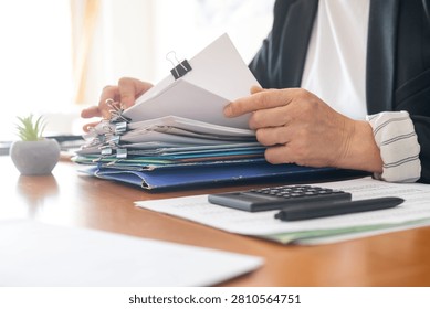 Person's hands organizing a stack of business documents, reports, and paperwork held together by binder clips on an office desk, illustrating administrative work and organization
