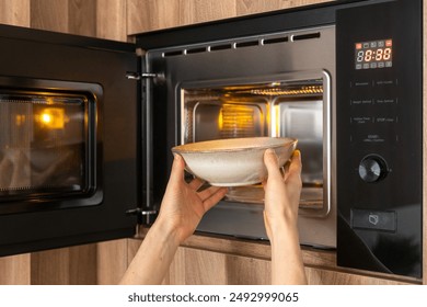 A person's hands are placing a bowl with soup into a microwave oven with the door open. Reheating food at home or at work during lunch