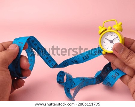 A person's hands pull a blue measuring tape wrapped around a small yellow alarm clock over pink.
