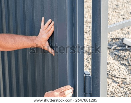 A person's hands are shown touching and adjusting a dark grey corrugated metal fence or gate panel next to a metal frame and hinge, with gravel on the ground.