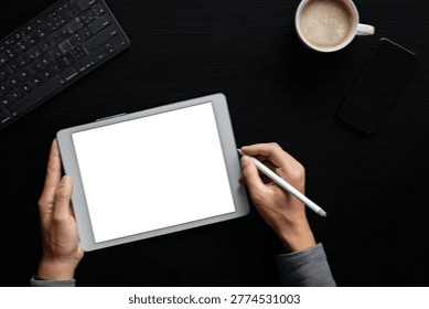 A person's hands are shown using a digital tablet with a stylus, next to a keyboard and a cup of coffee, on a dark surface.