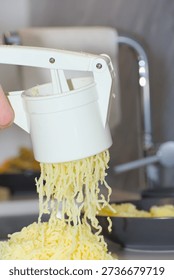 A person's hands use a white plastic manual potato ricer to press cooked yellow potatoes onto a pile of white flour on a kitchen countertop.