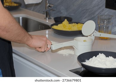 A person's hands use a white plastic manual potato ricer to press cooked yellow potatoes onto a pile of white flour on a kitchen countertop.