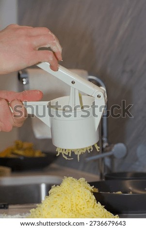 A person's hands use a white plastic manual potato ricer to press cooked yellow potatoes onto a pile of white flour on a kitchen countertop.