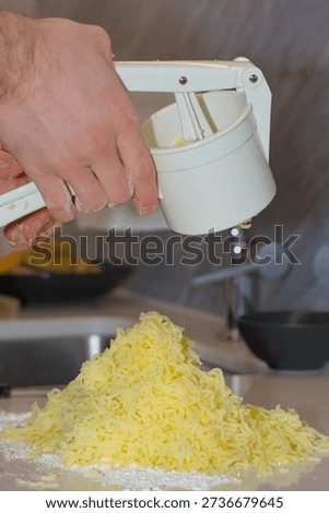 A person's hands use a white plastic manual potato ricer to press cooked yellow potatoes onto a pile of white flour on a kitchen countertop.