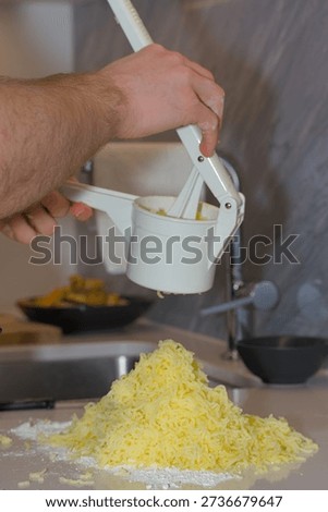 A person's hands use a white plastic manual potato ricer to press cooked yellow potatoes onto a pile of white flour on a kitchen countertop.