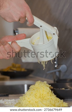 A person's hands use a white plastic manual potato ricer to press cooked yellow potatoes onto a pile of white flour on a kitchen countertop.