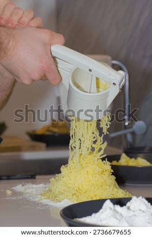 A person's hands use a white plastic manual potato ricer to press cooked yellow potatoes onto a pile of white flour on a kitchen countertop.