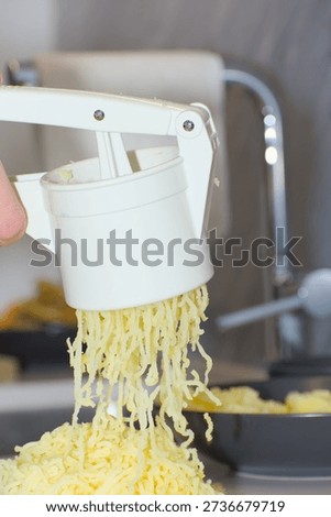 A person's hands use a white plastic manual potato ricer to press cooked yellow potatoes onto a pile of white flour on a kitchen countertop.