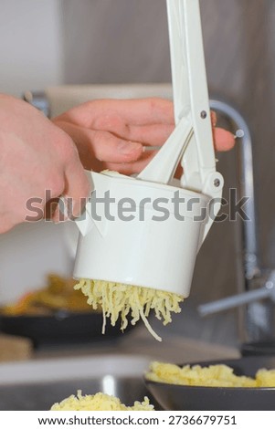 A person's hands use a white plastic manual potato ricer to press cooked yellow potatoes onto a pile of white flour on a kitchen countertop.