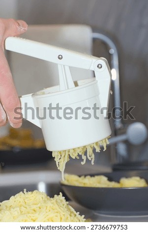 A person's hands use a white plastic manual potato ricer to press cooked yellow potatoes onto a pile of white flour on a kitchen countertop.