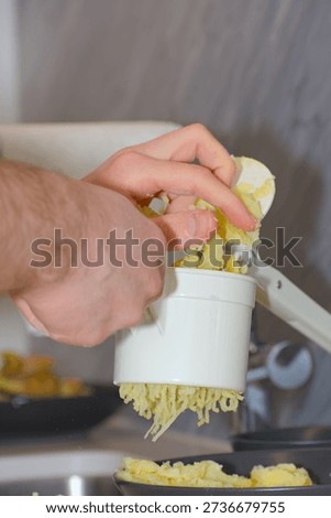A person's hands use a white plastic manual potato ricer to press cooked yellow potatoes onto a pile of white flour on a kitchen countertop.