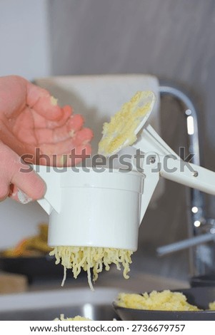 A person's hands use a white plastic manual potato ricer to press cooked yellow potatoes onto a pile of white flour on a kitchen countertop.
