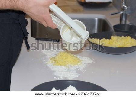 A person's hands use a white plastic manual potato ricer to press cooked yellow potatoes onto a pile of white flour on a kitchen countertop.