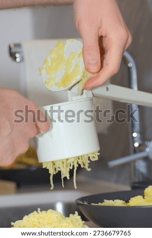 A person's hands use a white plastic manual potato ricer to press cooked yellow potatoes onto a pile of white flour on a kitchen countertop.