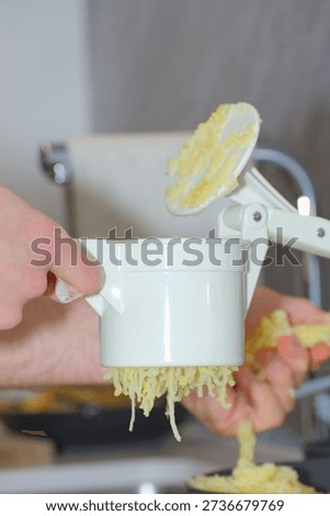 A person's hands use a white plastic manual potato ricer to press cooked yellow potatoes onto a pile of white flour on a kitchen countertop.