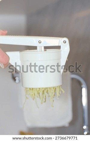 A person's hands use a white plastic manual potato ricer to press cooked yellow potatoes onto a pile of white flour on a kitchen countertop.