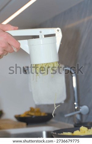 A person's hands use a white plastic manual potato ricer to press cooked yellow potatoes onto a pile of white flour on a kitchen countertop.