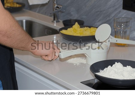 A person's hands use a white plastic manual potato ricer to press cooked yellow potatoes onto a pile of white flour on a kitchen countertop.