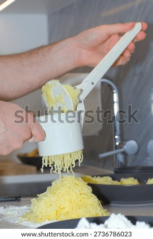 A person's hands use a white plastic manual potato ricer to press cooked yellow potatoes onto a pile of white flour on a kitchen countertop.