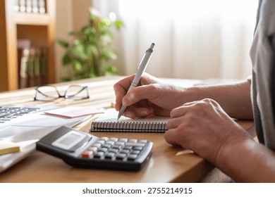 Person's hands using a calculator and writing notes in a notebook, managing finances, planning budget, and working on solutions for business operations at a desk