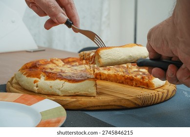 Person's hands using a fork and knife, lifting a delicious slice of hot homemade pizza from a wooden board