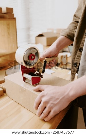 Persons hands using handheld dispenser for cutting tape on cardboard box in office setting, boxes and supplies visible on desk