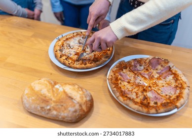 Person's hands using a knife and fork to cut a hot pizza into slices, ready for sharing a casual meal with friends or family gathered around a wooden table with another whole pizza and fresh bread